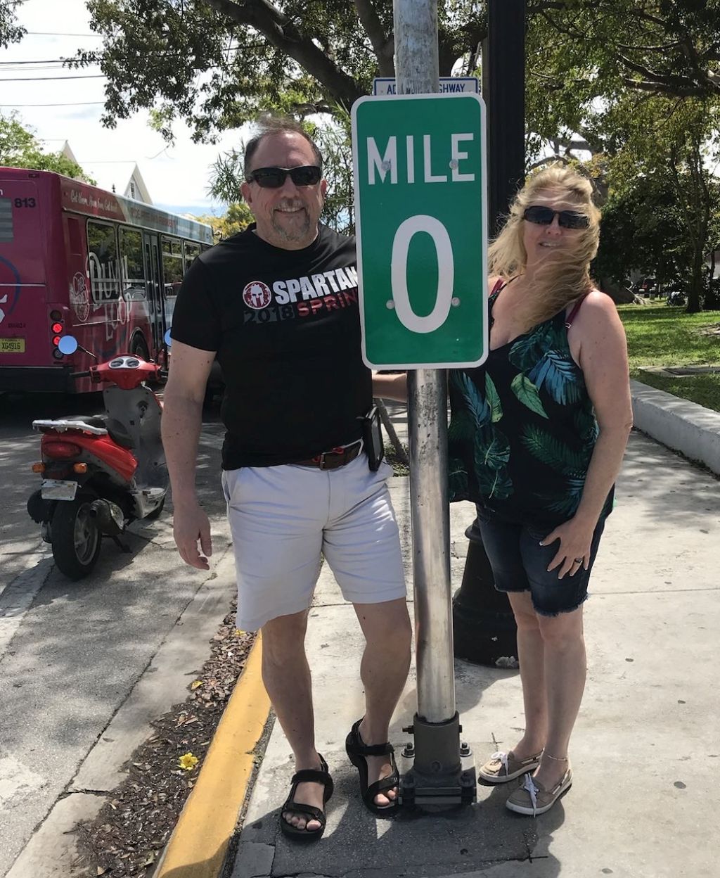 A couple wearing sunglasses pose on either side of the Mile 0 sign in Key West, Florida