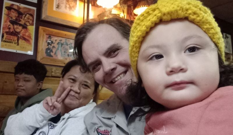 A Christian couple proudly shows off their daughter, who is in the forefront wearing a yellow toque in a restaurant