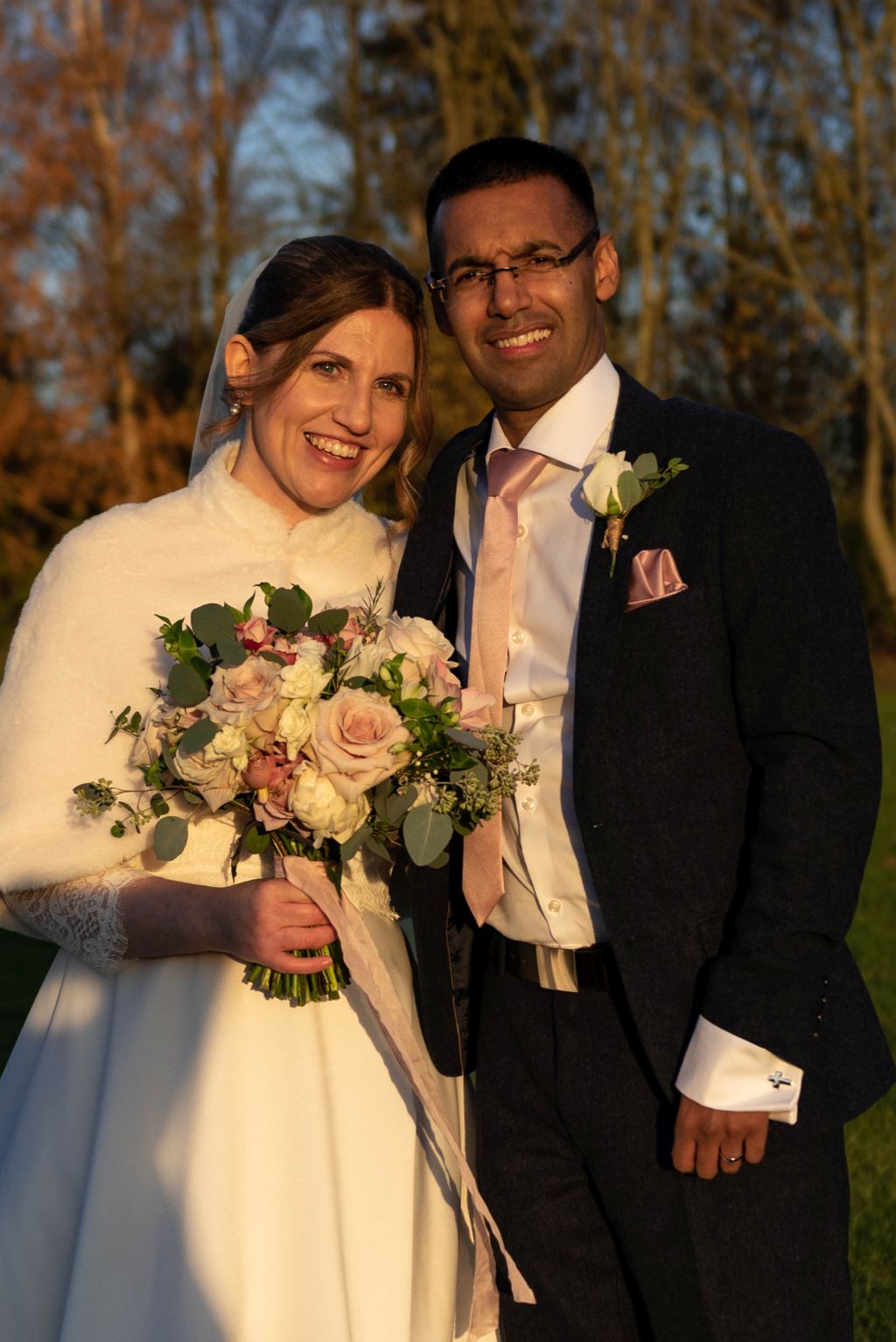 A beautiful White woman in a wedding dress is full of joy as she stands next to her new husband, a handsome Black man, as they pose outdoors on a sunny Autumn day