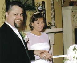 A man grins as he blindly pushes his wife's hand down as she tries cutting the cake