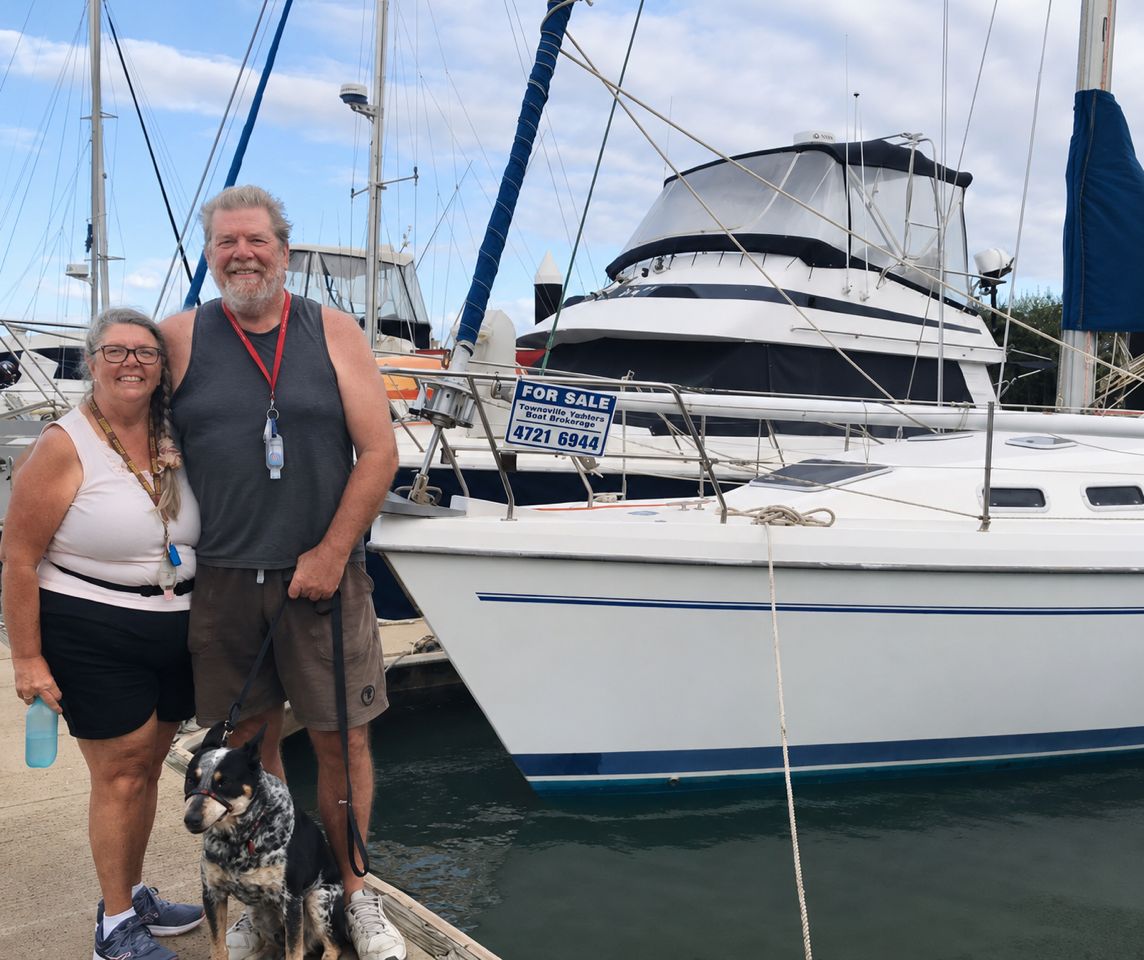 A married couple stand on a dock with their dog, next to a massive boat they have purchased