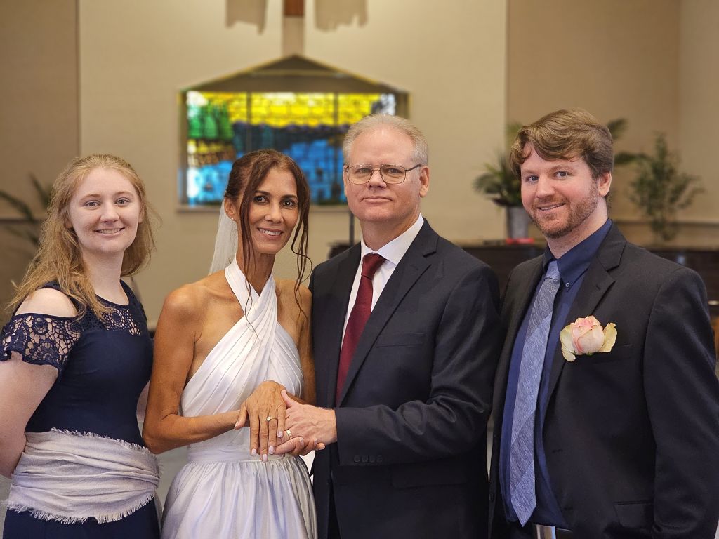A new bride in a sleeveless white dress stands with her new husband flanked by their wedding party, at church