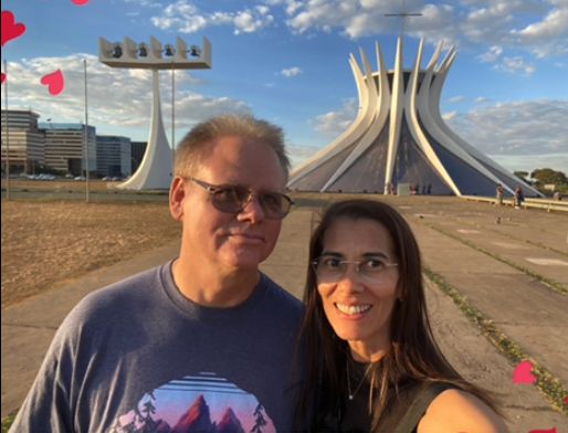 A Christian couple in glasses pose in the sun in front of the Cathedral of Brasília