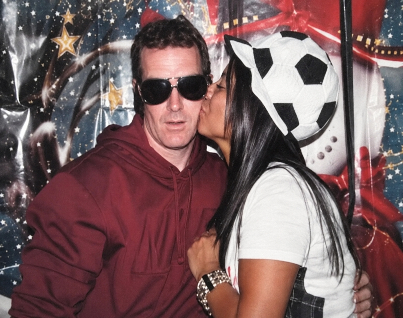 A dashing man in sunglasses is kissed lovingly by a woman in a white football hat