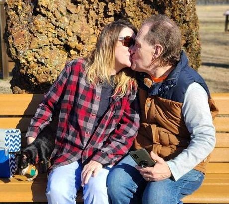 A woman in a plaid shirt kisses a man while they sit on a park bench with their dog