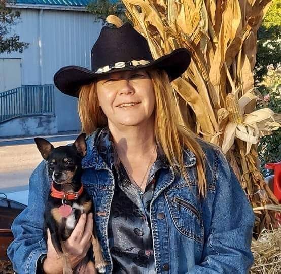 A pretty blonde woman in a black cowboy hat smiles while holding her dog at an Autumn festival