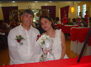 A man in glasses holds his new wife's hands on their wedding day. Both are dressed in white and all the tables have red and white covers