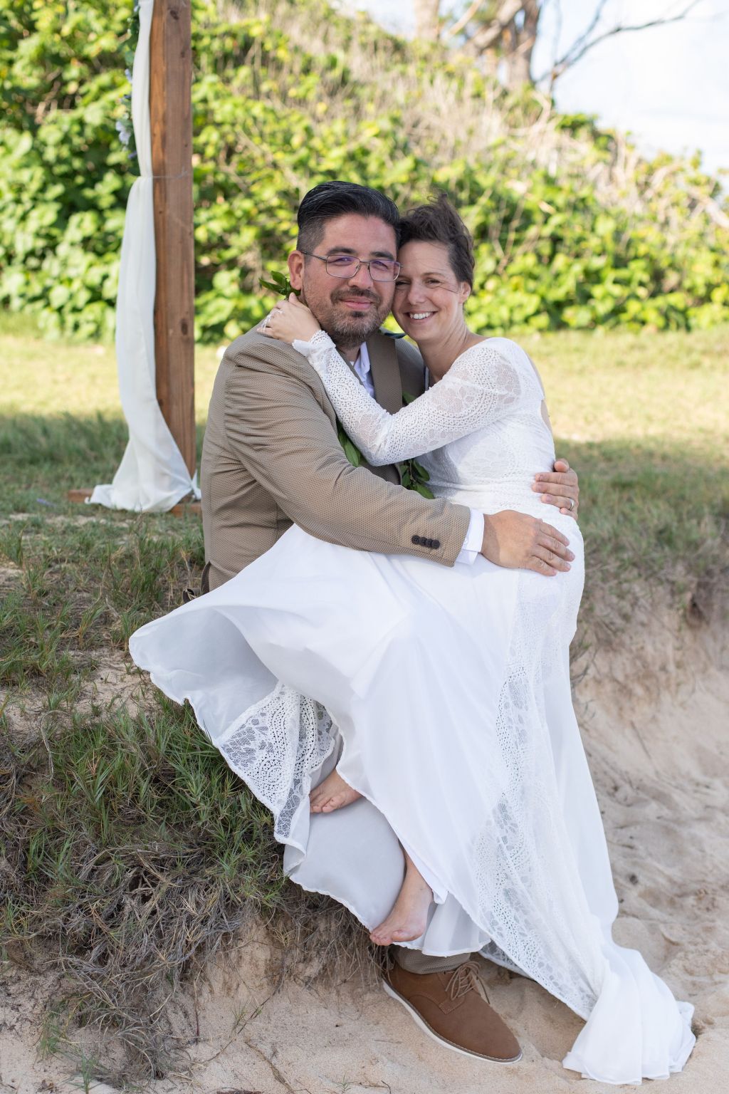 A barefoot bride leaps on to her sitting husband who catches her, as he sits on a bank by the sand.