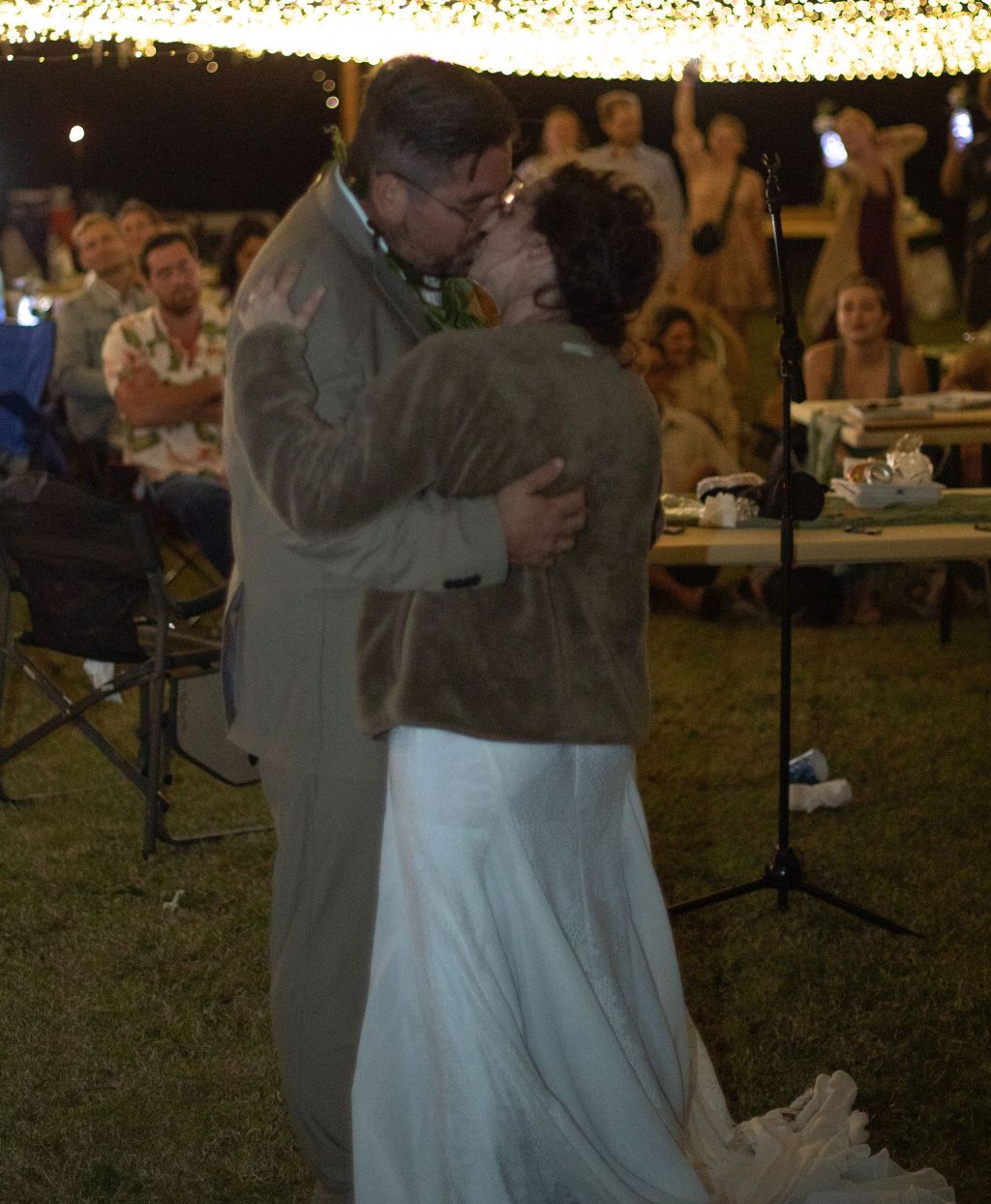 A husband kisses his new wife at an outdoor reception at night, surrounded by friends and family
