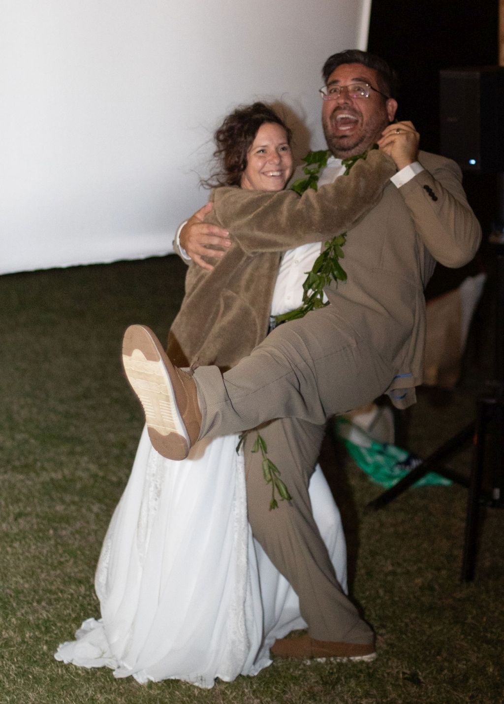 A husband in a brown suit does a silly dance outdoors while holding his laughing wife