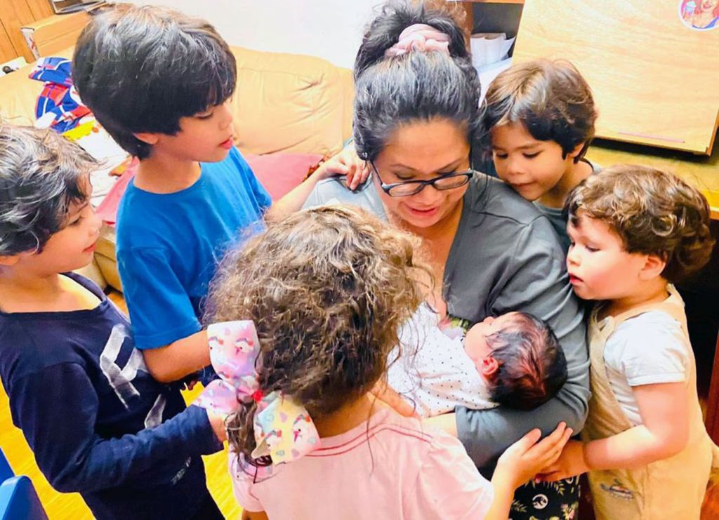 A mother cuddles her newborn at home, while surrounded by 5 curious children.