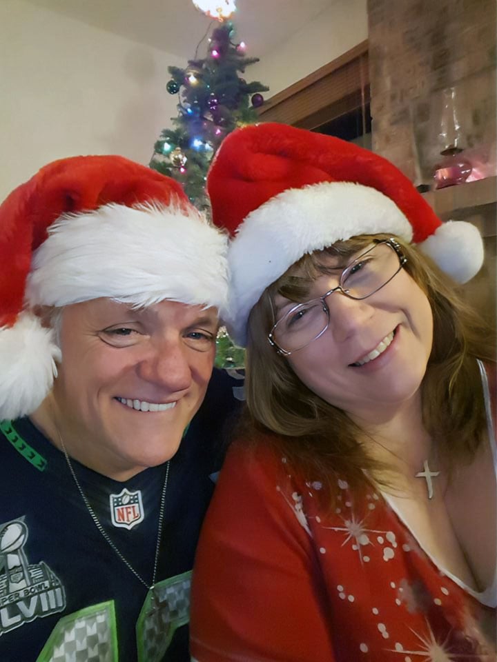 A mature Christian couple laugh as they pose for a Christmas photo together, while wearing Santa hats