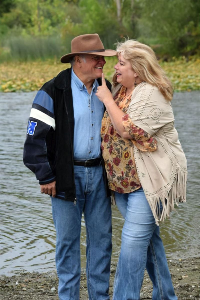 A woman affectionately touches her husband's cheek as they stand by the water on a cool, windy day