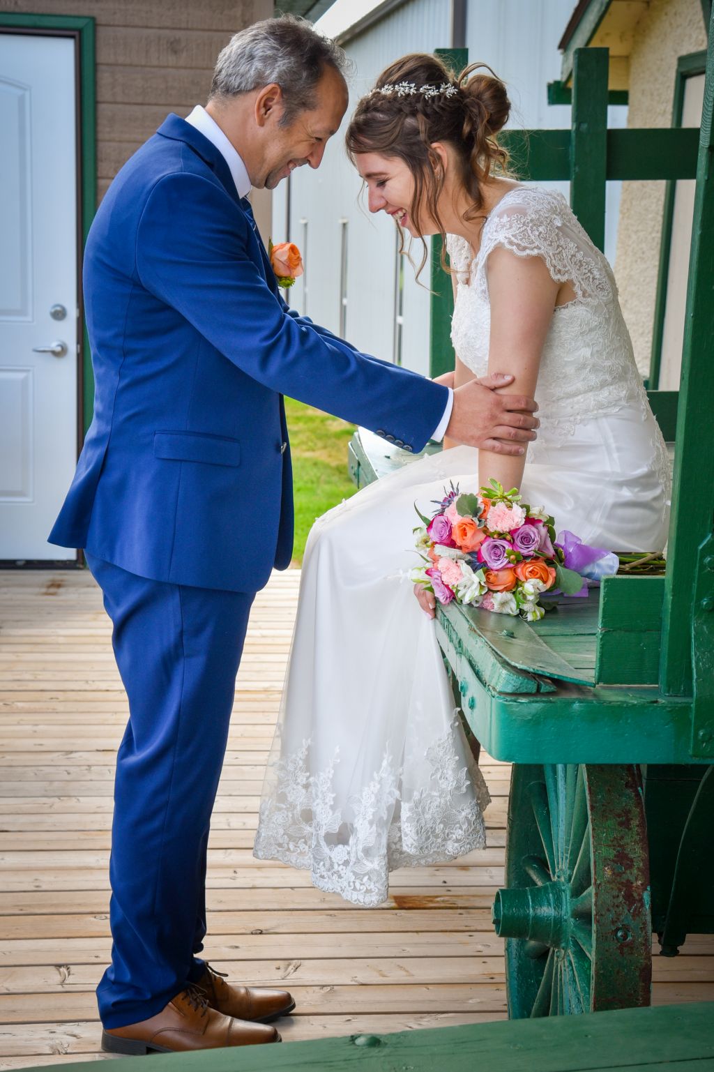 A groom in a navy blue suit shares a joke with his beautiful bride, while she is seated outdoors on a pioneer cart