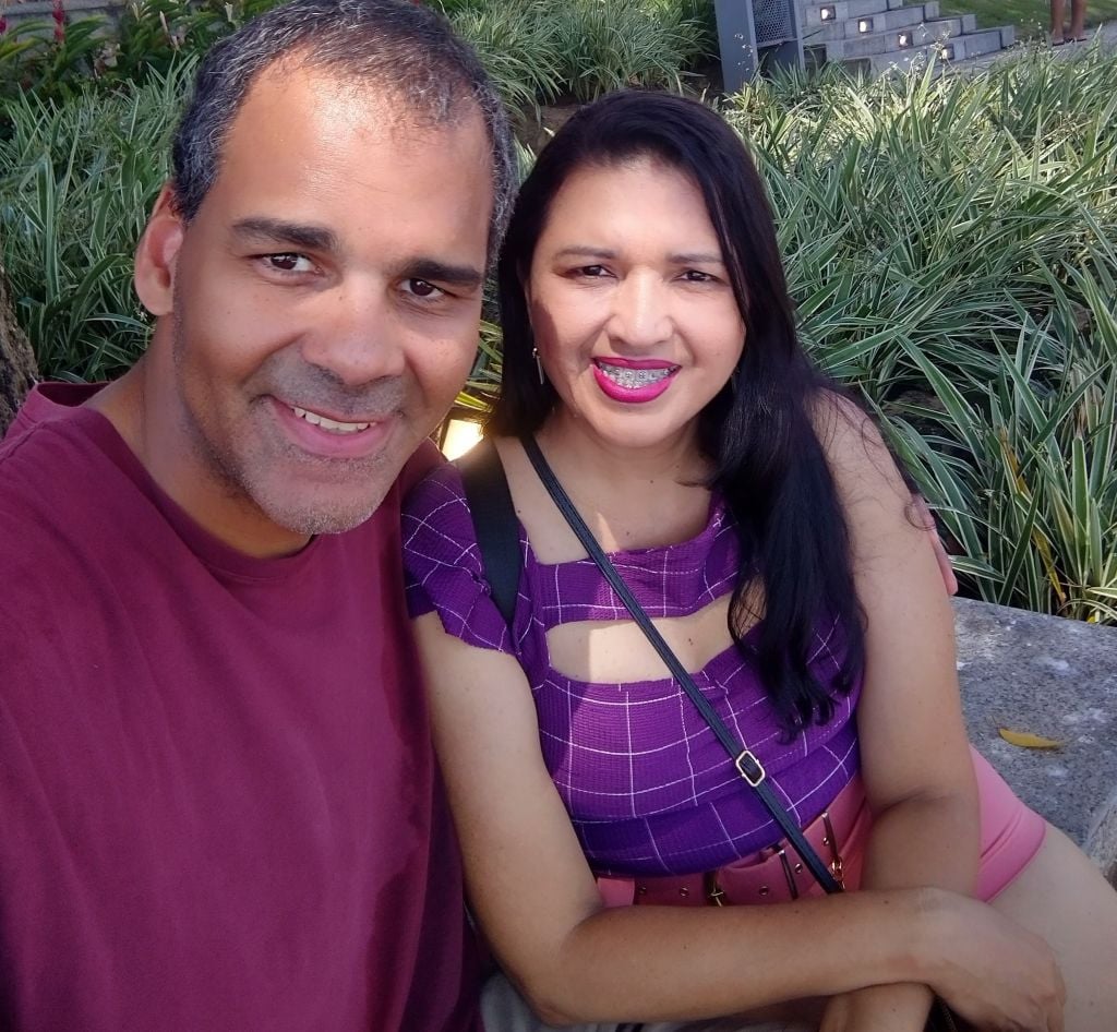 A Brazilian Christian woman smiles next to a man who has his arm around her while they pose for a selfie in a garden