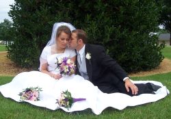 A beautiful bride sits on the grass with her husband who kisses her