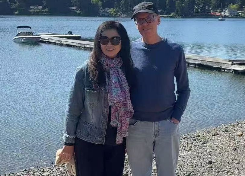 A woman in sunglasses and jean jacket stands on a pebble beach next to a man in a hat and glasses, in front of a beautiful lake