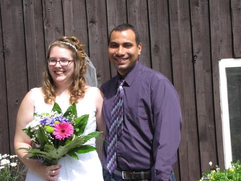 An elated Christian couple stand with bouquet in front of pioneer building