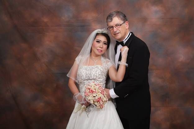 Christian senior marries Indonesian bride who tenderly holds onto him in her white wedding dress