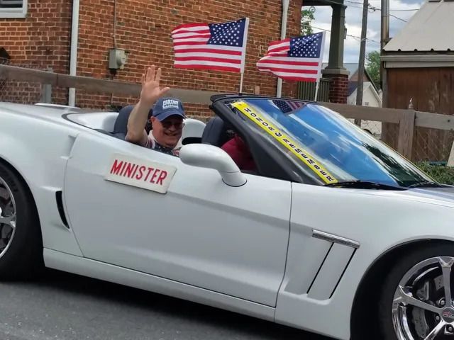 A man seated in a white sports car with American flags waves and smiles at the camera
