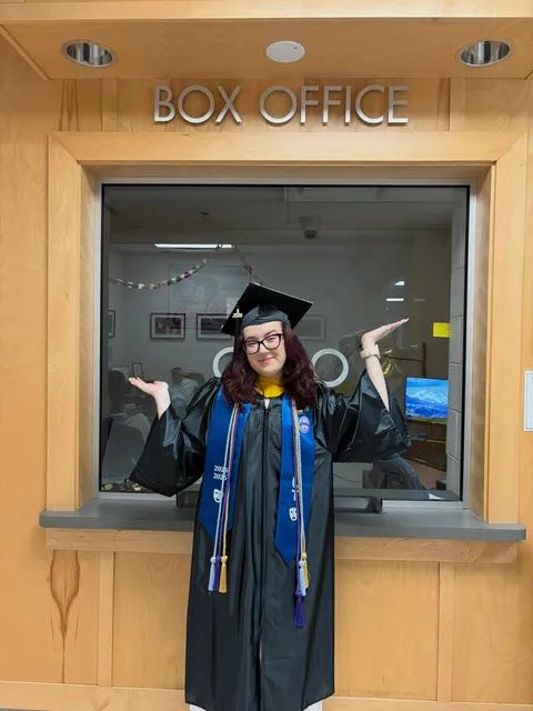 A young woman in graduation dress poses in front of a Box Office booth