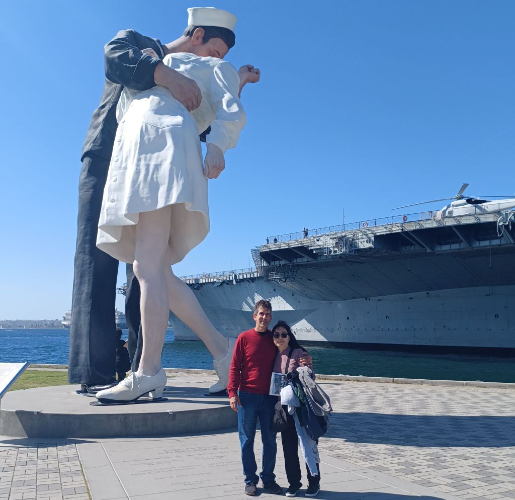 A newly reunited couple pose in front of The Embracing Peace statue and USS Midway in San Diego