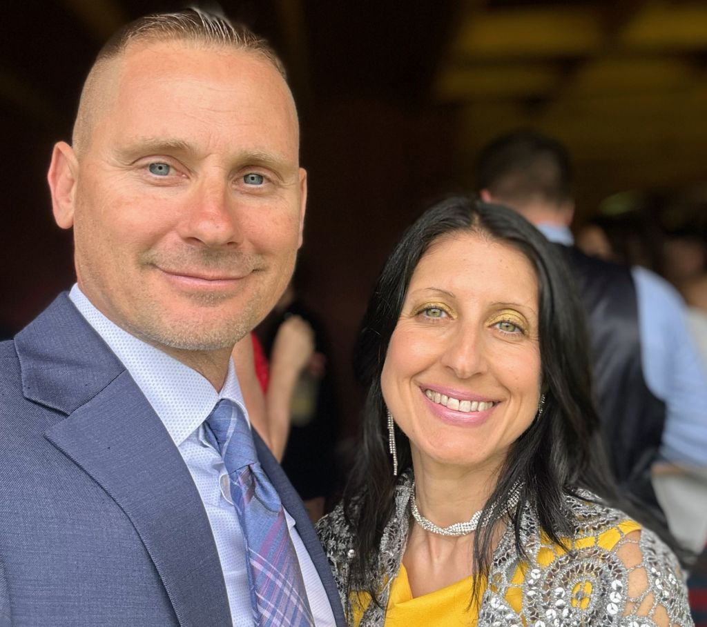 A man wearing a blue-grey suit and a with short hair poses next to a woman in a yellow dress