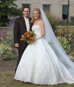 A beautiful woman in a wedding dress with a long veil stands next to her husband and holds her bouquet