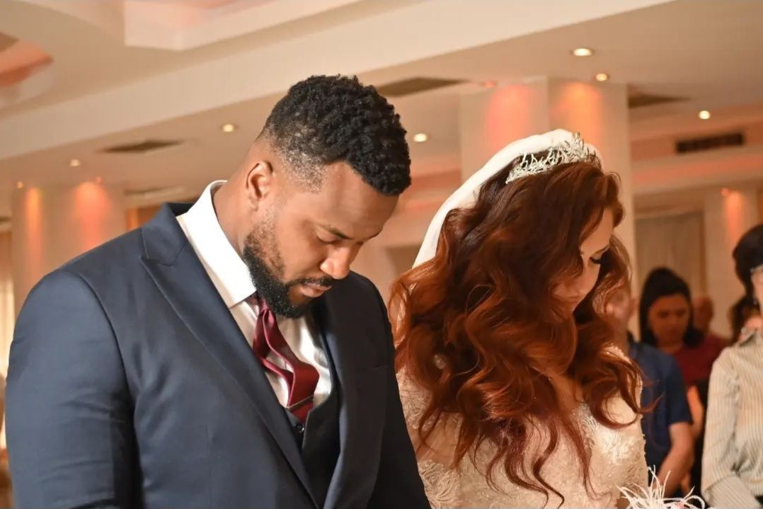 A well groomed Black man in a suit bows his head in prayer as he stands next to his White wife, as they receive a blessing on their wedding day.