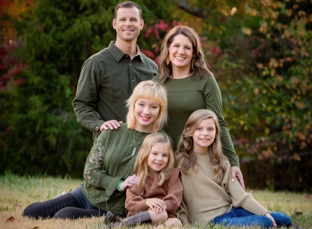 A couple celebrating their 15th anniversary pose with their daughters outdoors in front of a beautiful copse