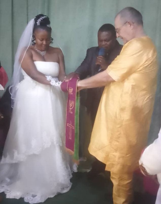 A beautiful African woman in a white wedding dress marries a White man in a gold outfit as a pastor prays over them.