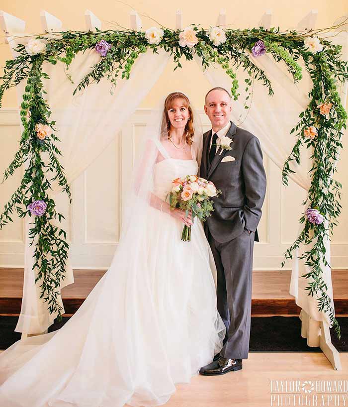 A couple smile under an arch with flowers