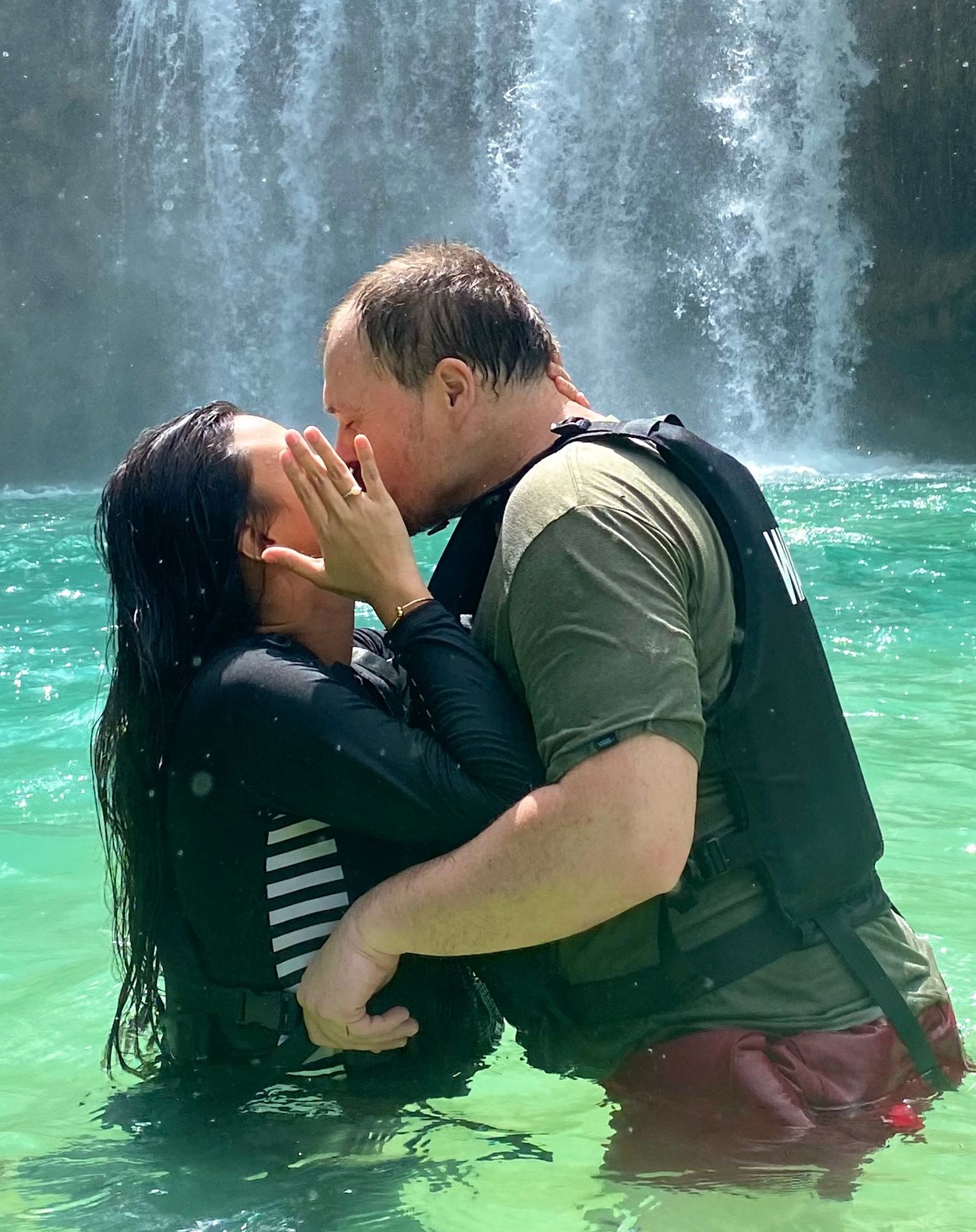 A woman in front of a waterfall shows off her ring as a man leans over to kiss her.