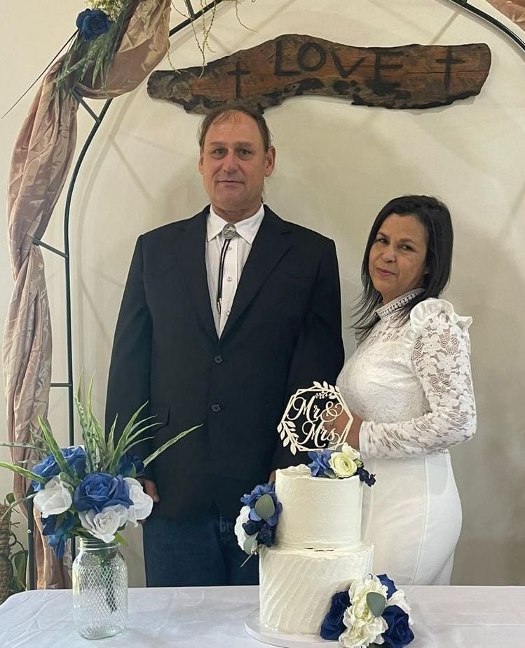 A newly married mature couple pose in front of a wedding cake under a sign which says LOVE. The groom is wearing black, while the bride is wearing a white dress.