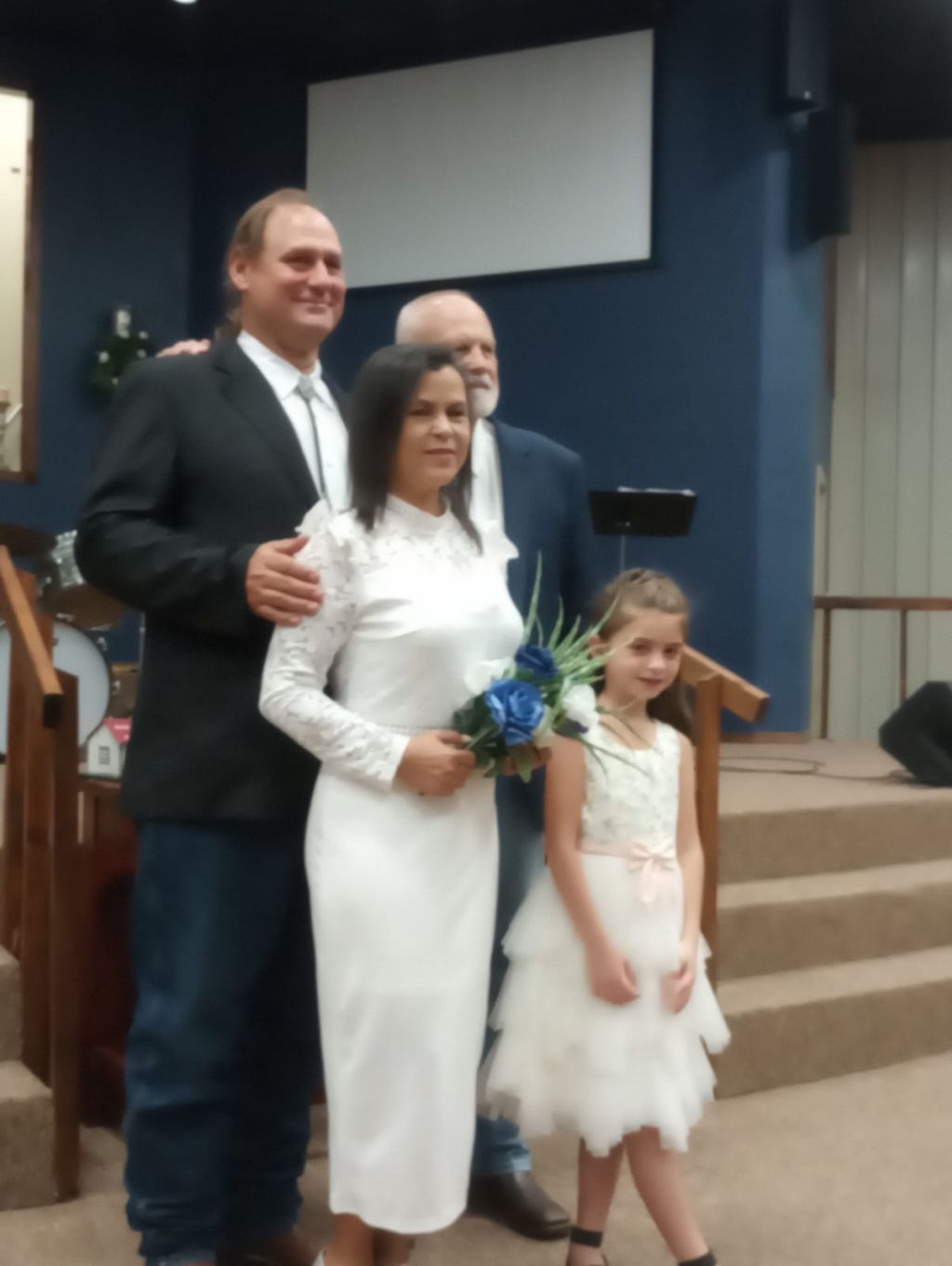 A man beams with pride as he stands behind a pretty woman in a form fitting wedding dress as she holds a small bouquet of flowers, while both stand in front of a church next to a flower girl