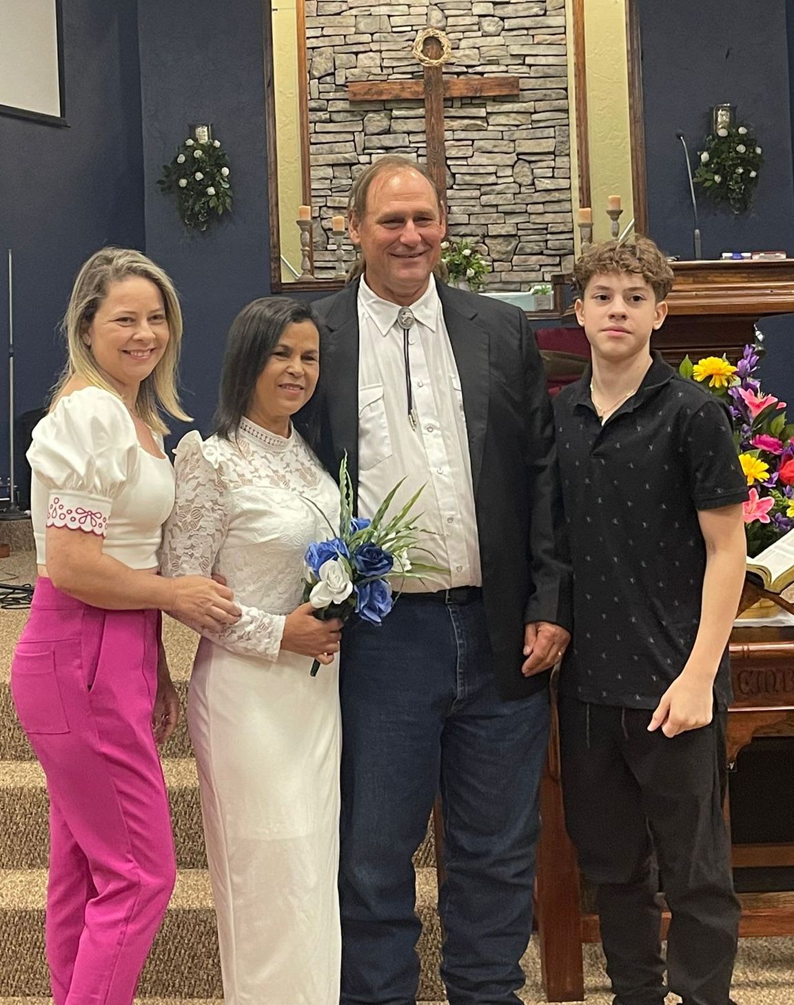 A tall American groom in a black sports jacket stands next to a beautiful woman in a white wedding dress. They are flanked by a young blonde woman and a smartly dressed teenaged boy as they pose in front of a church