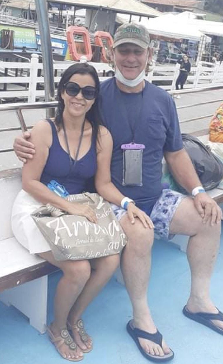 A smiling man in a baseball cap with COVID mask hanging below his chin grins as he has his arm around a beautiful Brazilian woman as they both sit on a tour boat which is docked