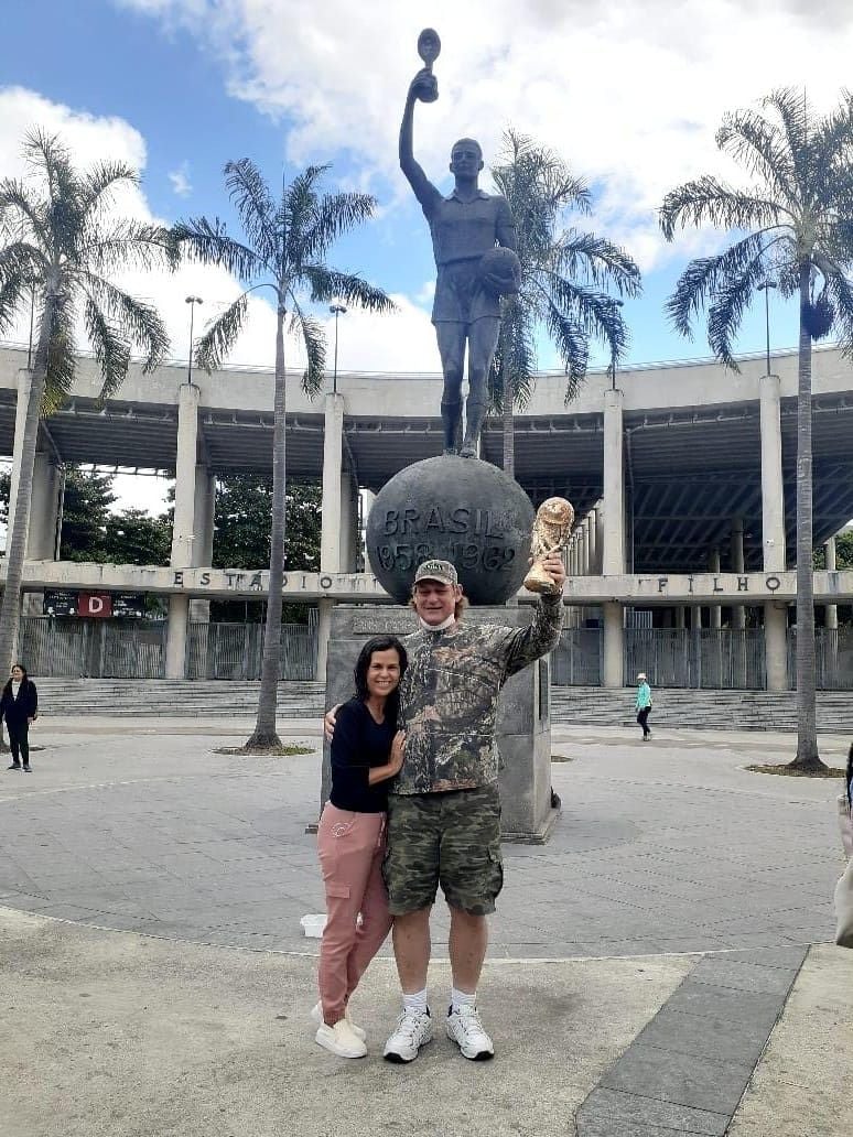 An American Christian man holds up a replica Fifa World Cup trophy in front of Estatua do Bellini in Rio de Janeiro, while his Brazilian wife smiles and hugs him