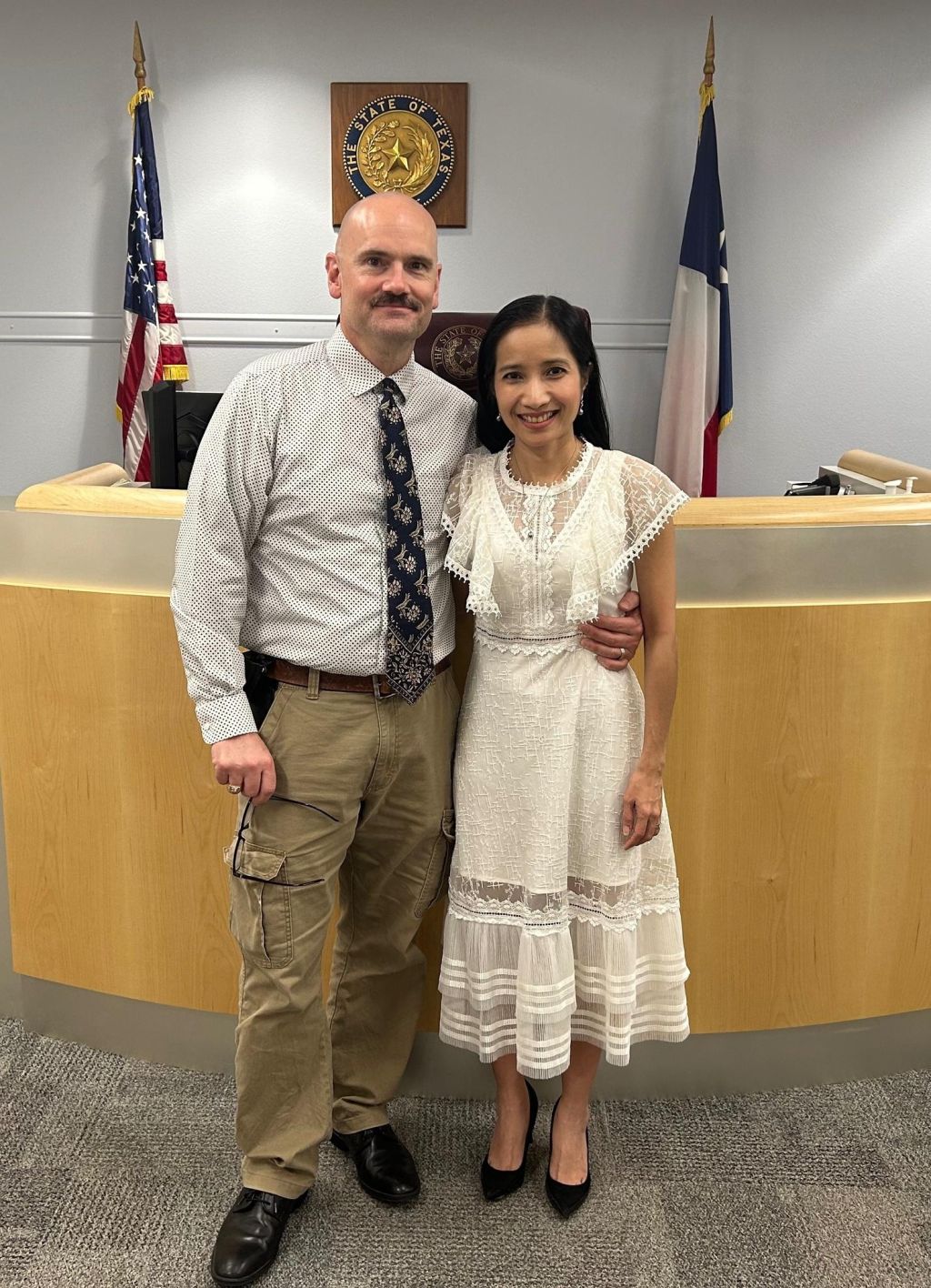 A man sporting a moustache and tie has his arm around a pretty woman in a nice white dress at the state of Texas registry