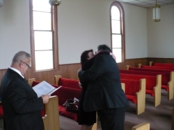 A pastor shyly looks away as a newly married couple wholeheartedly embrace