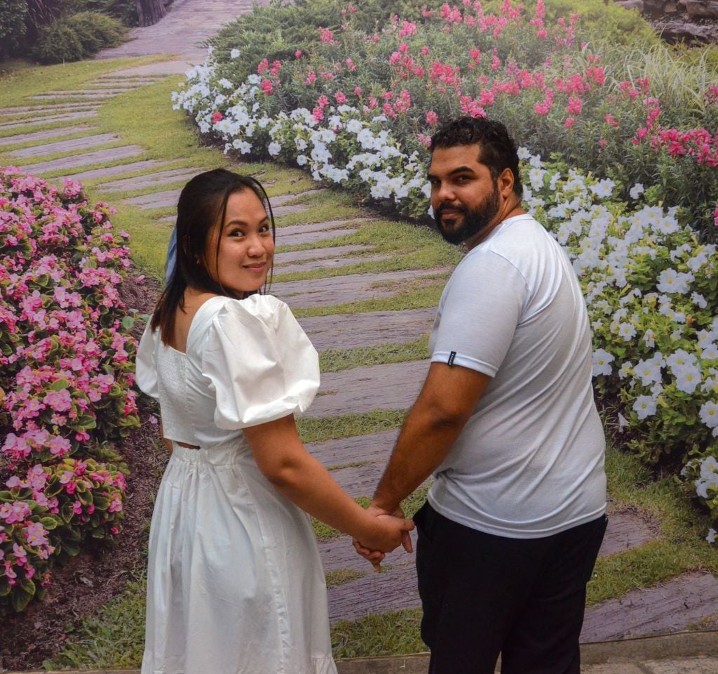 A young Christian couple holds hands while walking on a flower lined path, and looking back at the camera