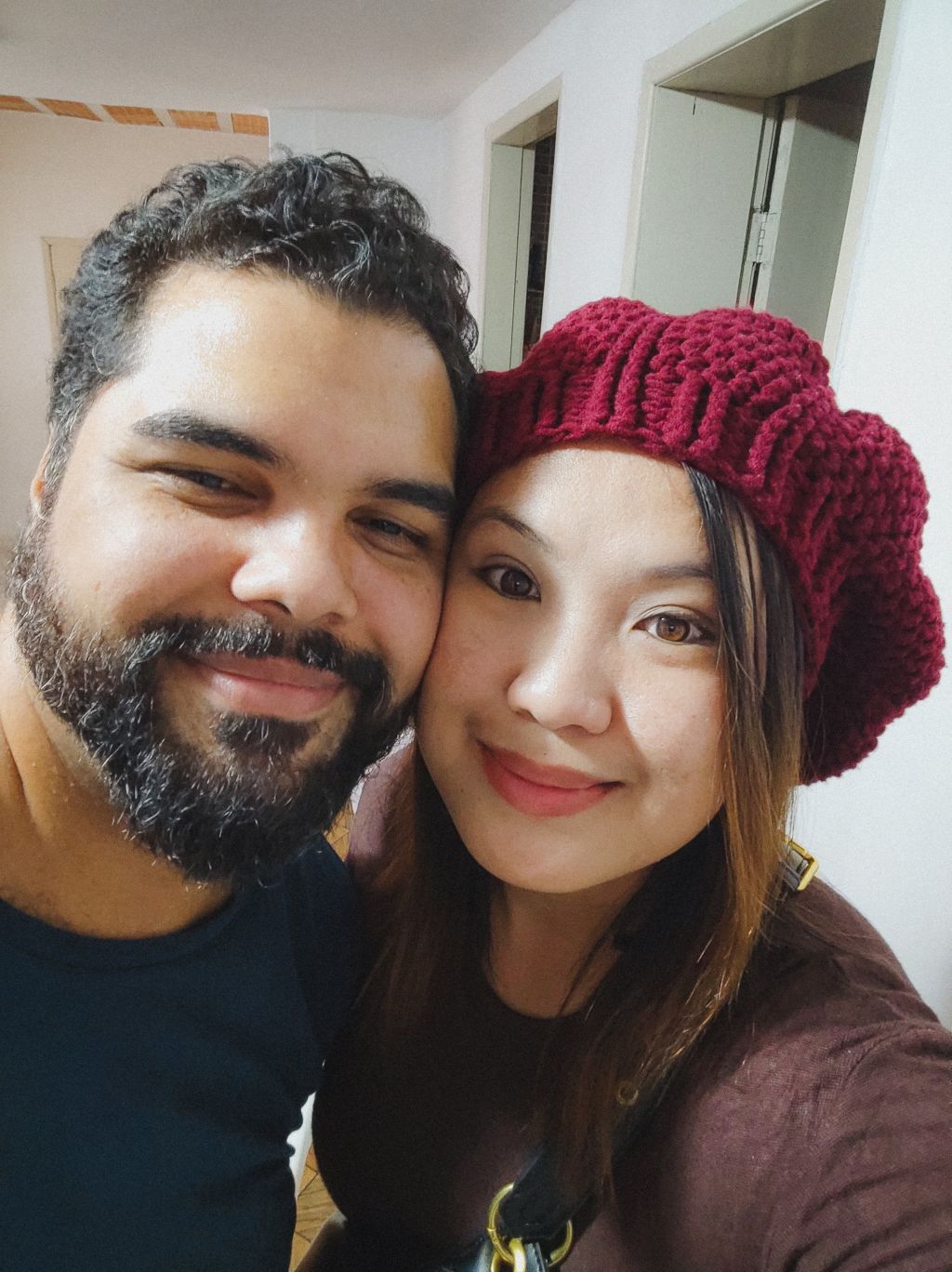 A Christian Filipina woman in a red beanie smiles cheek to cheek with a well groomed Brazilian man