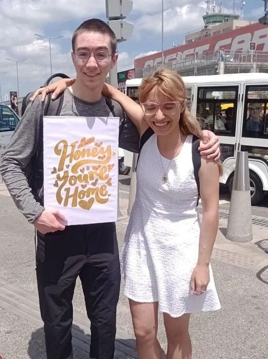 A young Christian man holds a sign at a bus stop which says Honey Youre Home as a woman holding him bursts out laughing