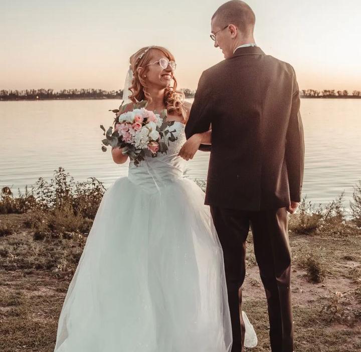 A young Christian bride gazes lovingly at her new husband as they face each other, arms linked, by the water