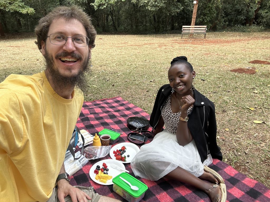 A bearded Caucasian man laughs while taking a selfie at a picnic with a pretty Black woman who is seated on the picnic blanket while smiling