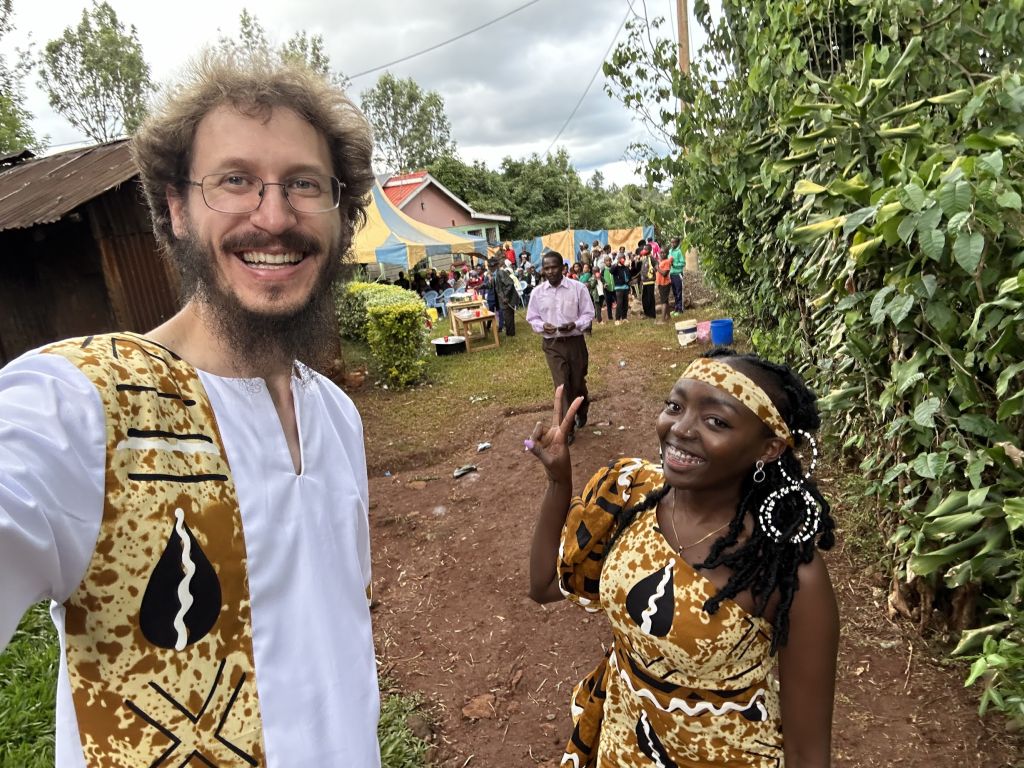 A man wearing a white tunic and gold vest smiles next to a pretty woman wearing a gold, brown and black patterned dress and headband while giving the peace sign at an outdoor festival in Kenya