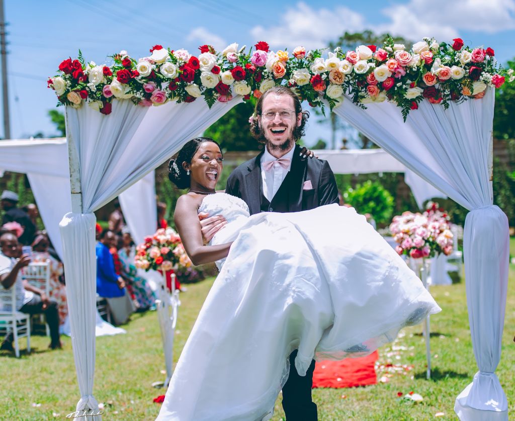 A newly married Christian groom laughs as he carries his new bride in her white wedding dress across a threshold adorned with flowers at an outdoor wedding