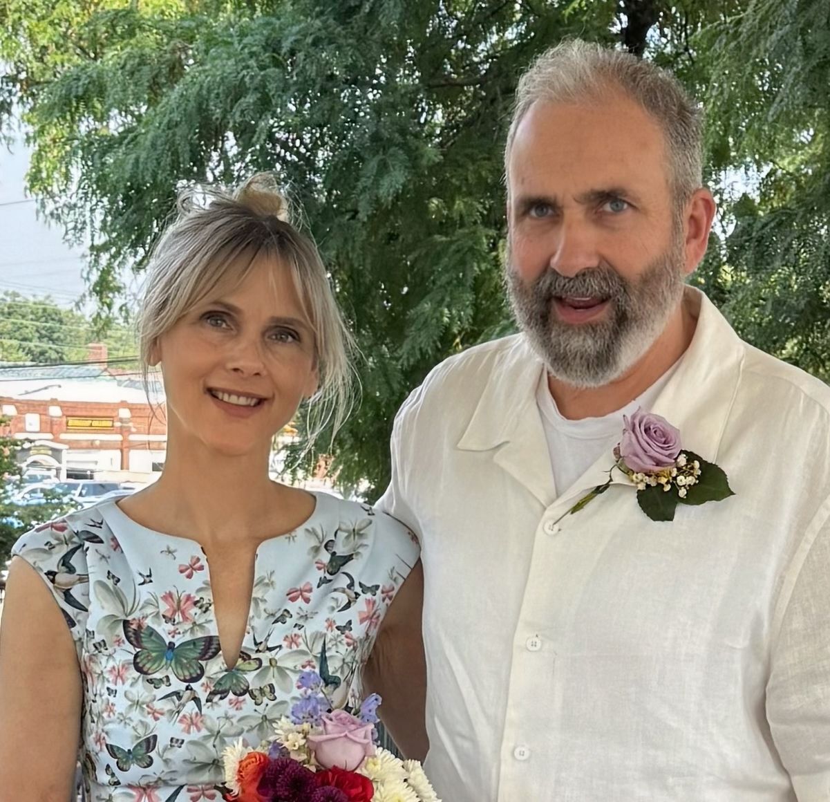 A pretty woman in a white dress holds a bouquet of flowers while a pleased man in a beard poses next to her