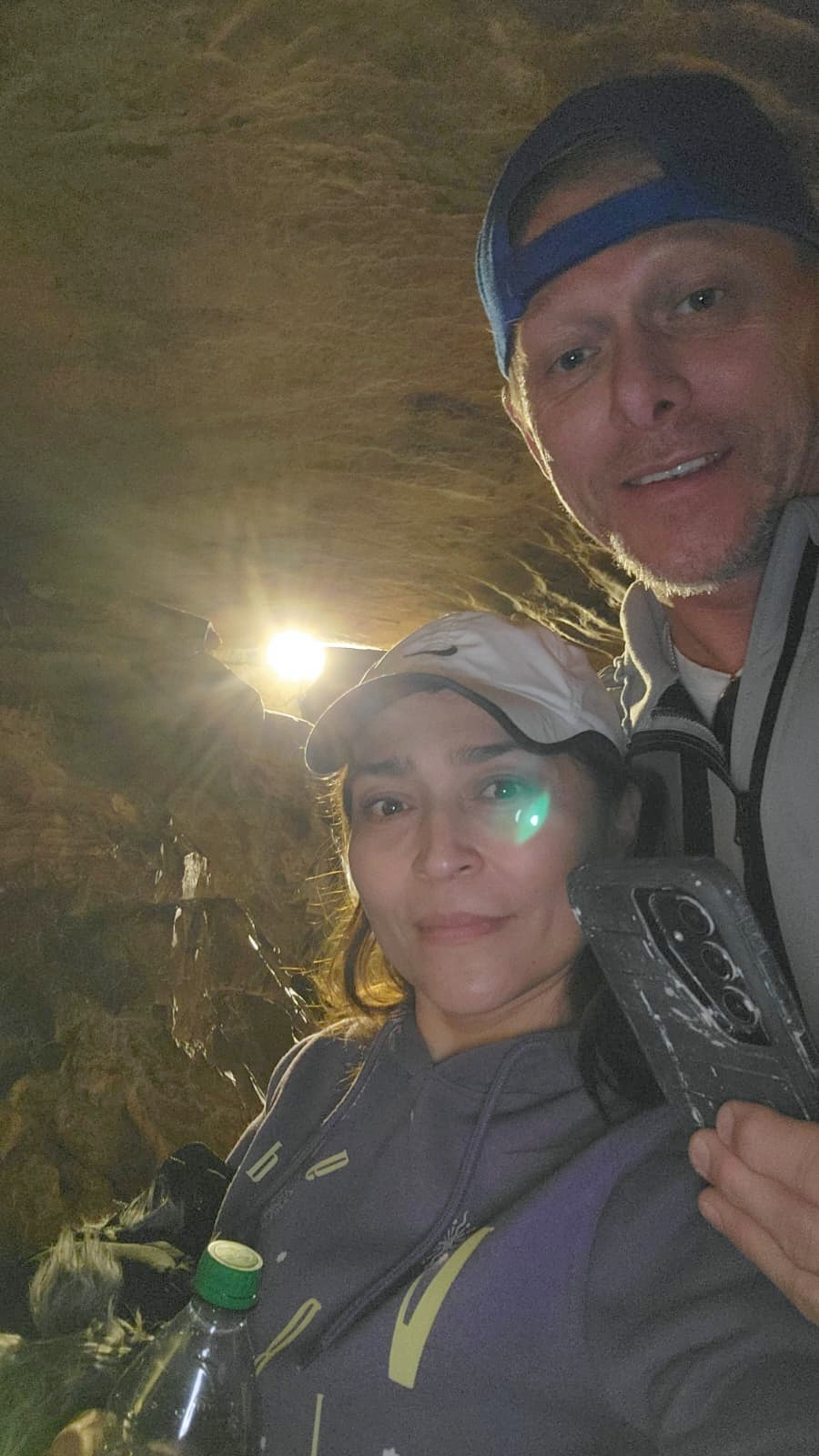A man in a baseball cap and jacket stands above a woman holding a water bottle as the 2 of them stand in a cave with a bright light in the background