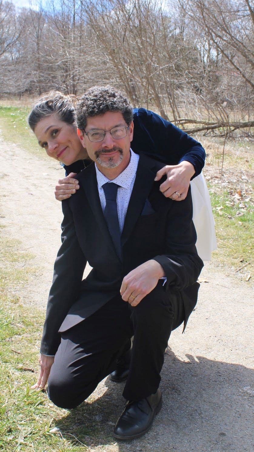 A woman peaks from behind a man in a suit, who is kneeling on a forest path